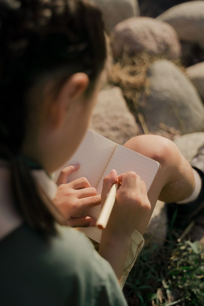A girl in scout uniform writes in a notebook while sitting on rocks outdoors.